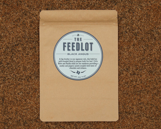 A brown paper bag with a round blue and white label reading "The Feedlot Black Angus" on a brown speckled surface. The label details the product, "Black Angus" by The Feedlot, as a bold and well-rounded black Angus blend with notes of chocolate, molasses, and Pasilla chilis, weighing 250 grams.