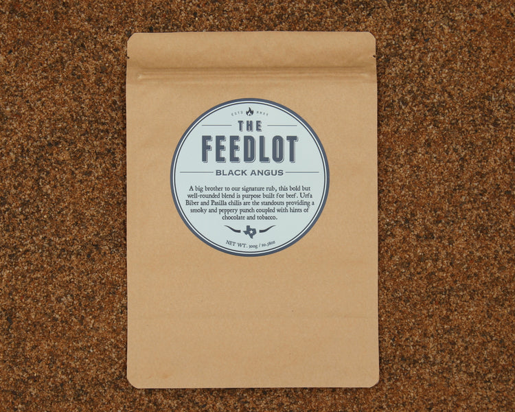 A brown paper bag with a round blue and white label reading "The Feedlot Black Angus" on a brown speckled surface. The label details the product, "Black Angus" by The Feedlot, as a bold and well-rounded black Angus blend with notes of chocolate, molasses, and Pasilla chilis, weighing 250 grams.