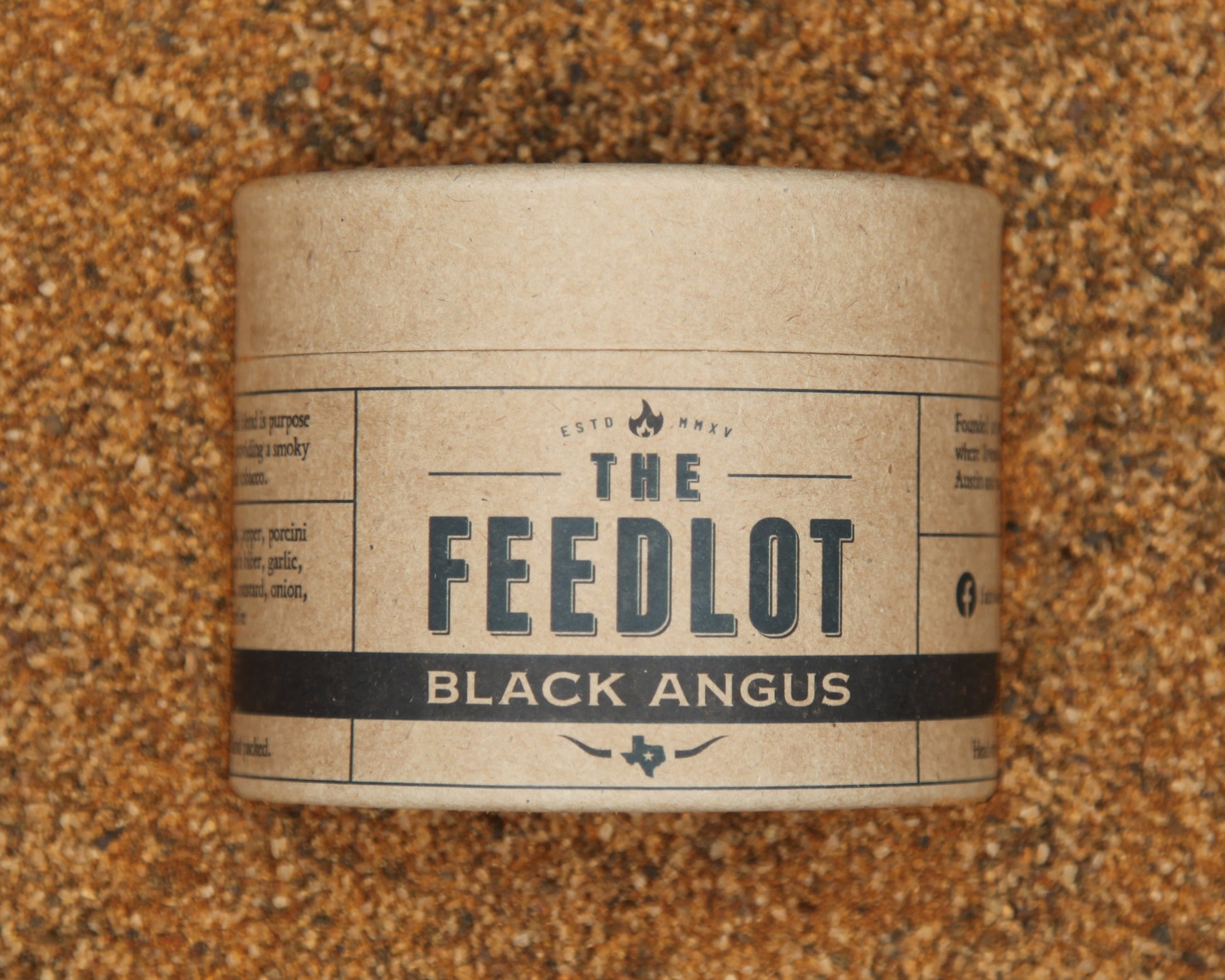 A cylindrical container labeled "The Feedlot Black Angus" is placed against a textured brown background. The label features various descriptive texts and icons, including a Texas state outline, indicating the product's branding as Black Angus by The Feedlot with a smoky and peppery punch.