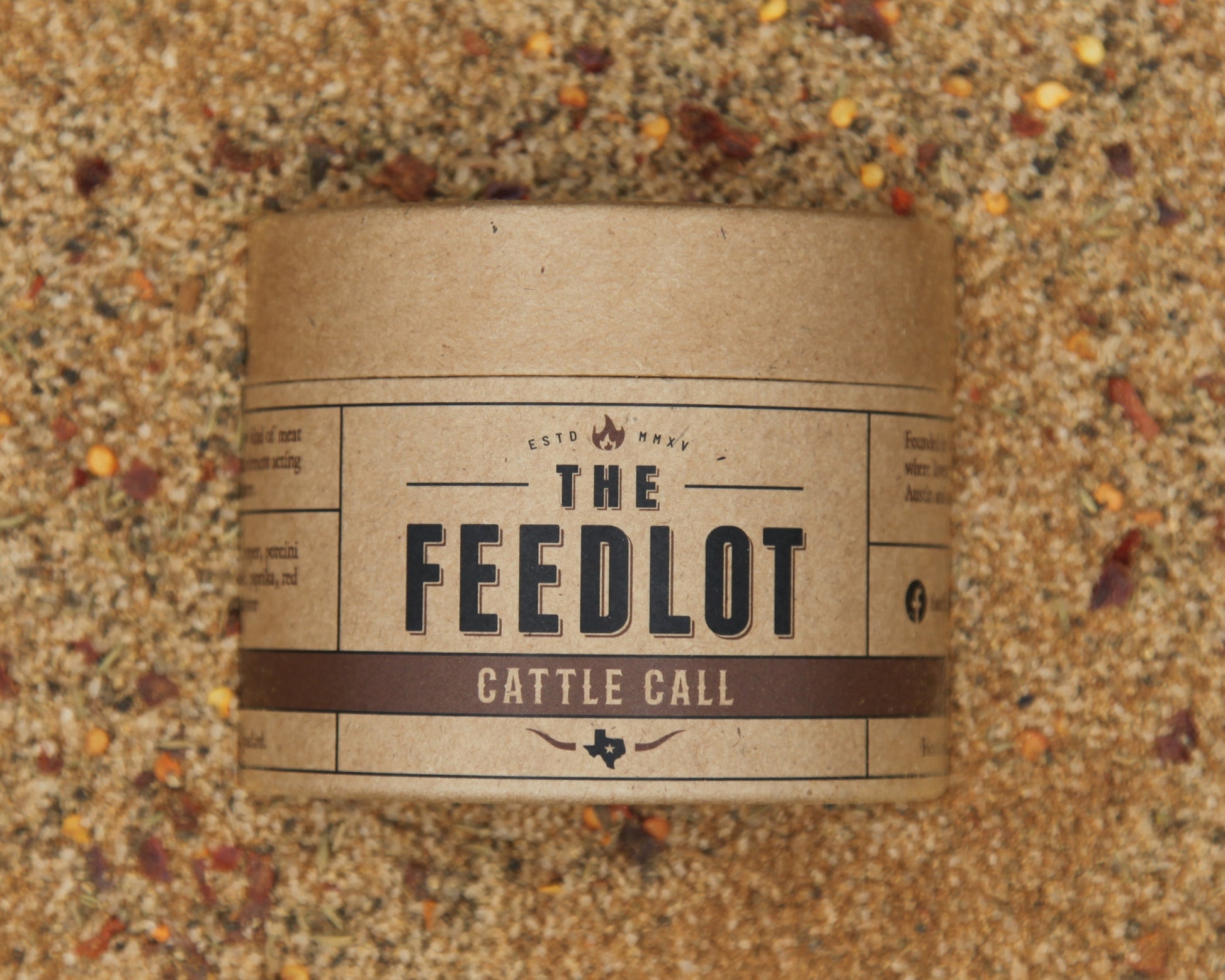 A cylindrical cardboard container labeled "The Feedlot Cattle Call" is placed on a bed of coarse, textured feed material. The label features a rustic design with brown and beige tones. Red pepper flakes and porcini mushrooms are scattered across the feed in the background.
