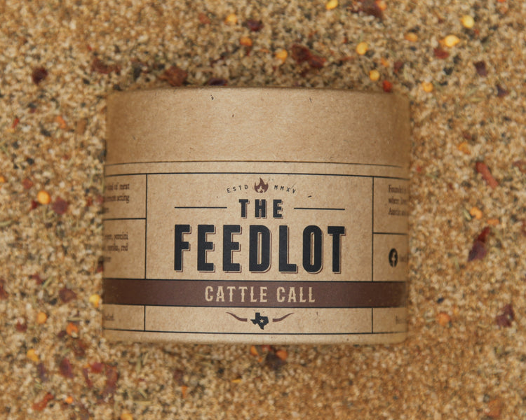 A cylindrical cardboard container labeled "The Feedlot Cattle Call" is placed on a bed of coarse, textured feed material. The label features a rustic design with brown and beige tones. Red pepper flakes and porcini mushrooms are scattered across the feed in the background.