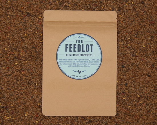 A brown paper package labeled "Crossbreed" by The Feedlot rests on a textured surface with mixed grains. The label touts a hybrid rub combining their signature blend, Carre Cali, with their renowned Black Angus rub. The net weight is 5.9 oz/160g.