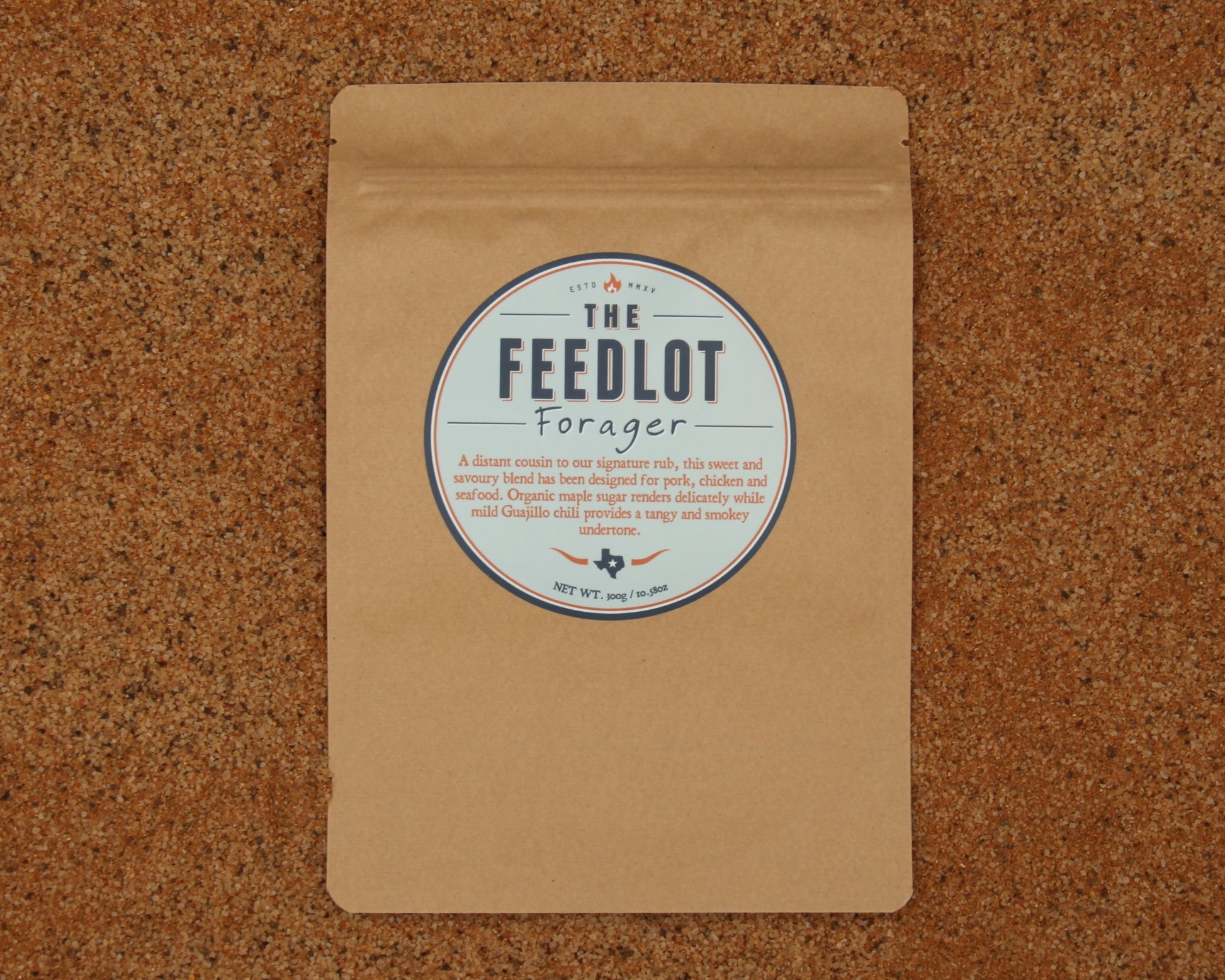A brown paper package with a circular label reading “The Feedlot Forager.” The label describes the product as *Forager* by *The Feedlot*, a mild organic coffee sweetened with organic maple sugar and perfectly roasted for poultry, pork, and meaty dishes. The package is placed on a cork surface.