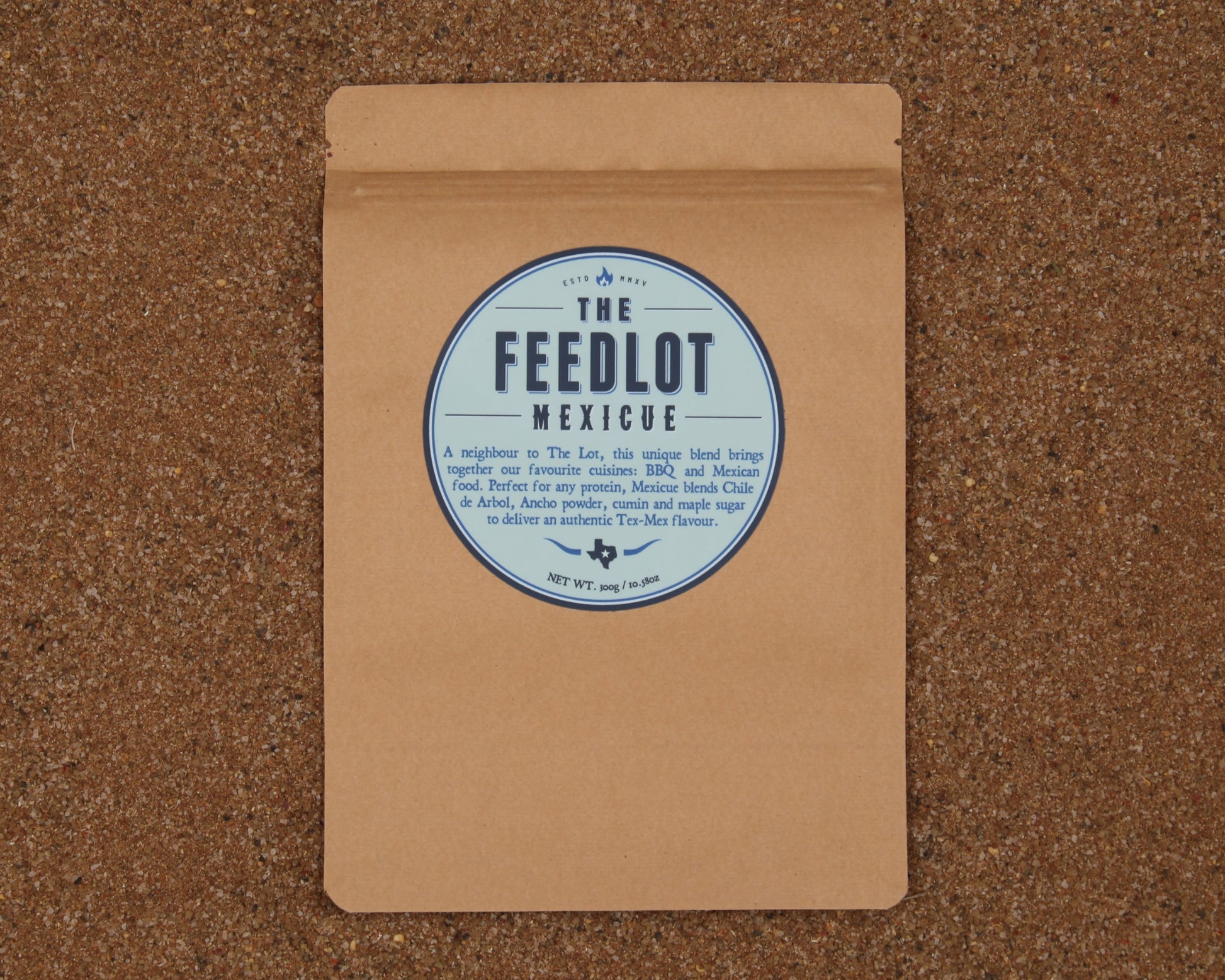 A brown paper package with a circular label that reads "The Feedlot Mexicue." The package is set against a textured, brown background. The label mentions that it contains a unique blend of spices for BBQ and Mexican cuisine, offering an authentic Tex-Mex flavor, weighing 75g.