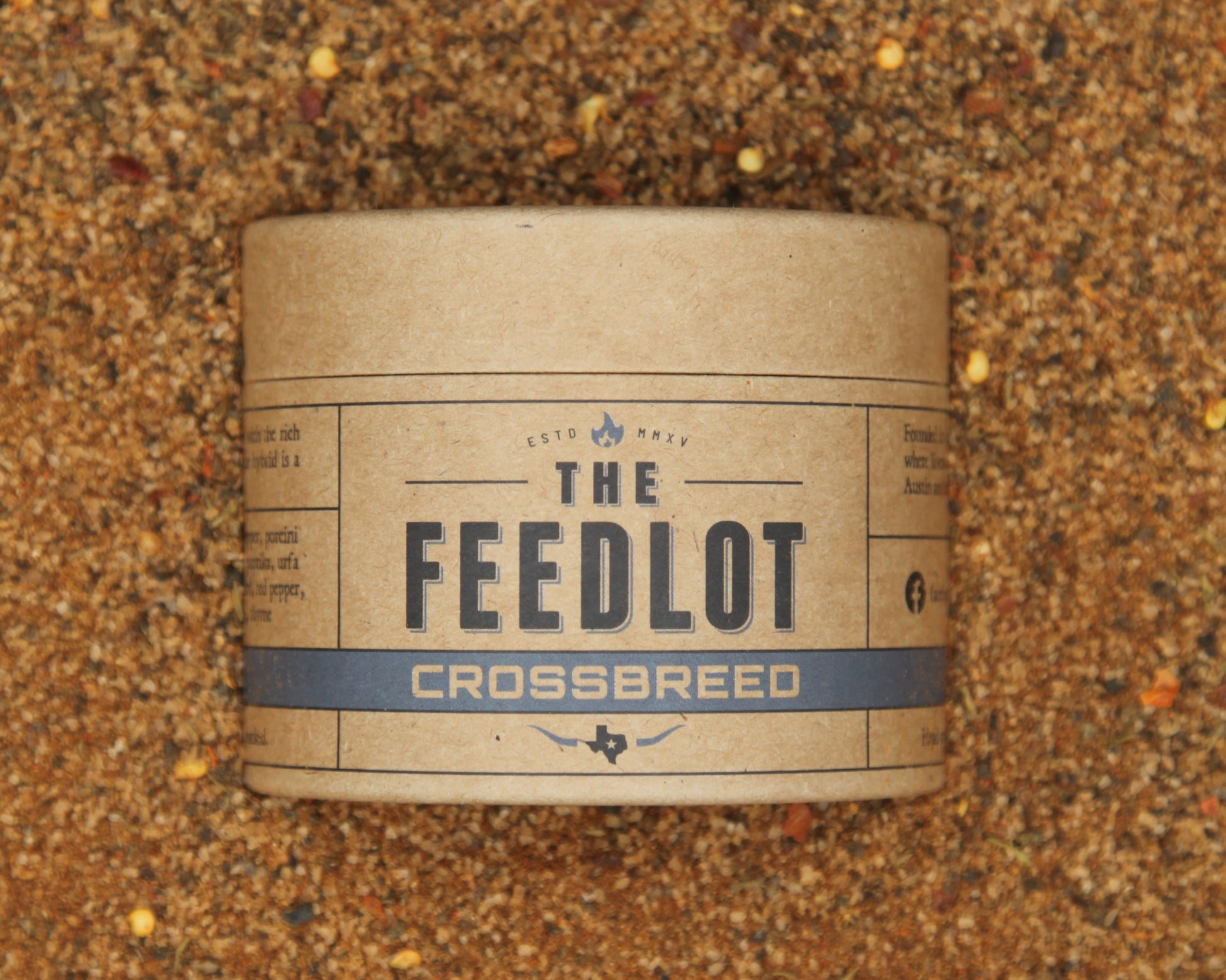 A round brown paper container labeled "Crossbreed" by The Feedlot sits on a textured sandy surface. The container features a simple, rustic design with a stripe and small icon, suggesting a natural product theme. This blend is perfect for your Black Angus cuts, enhancing the flavor like the ultimate rub.