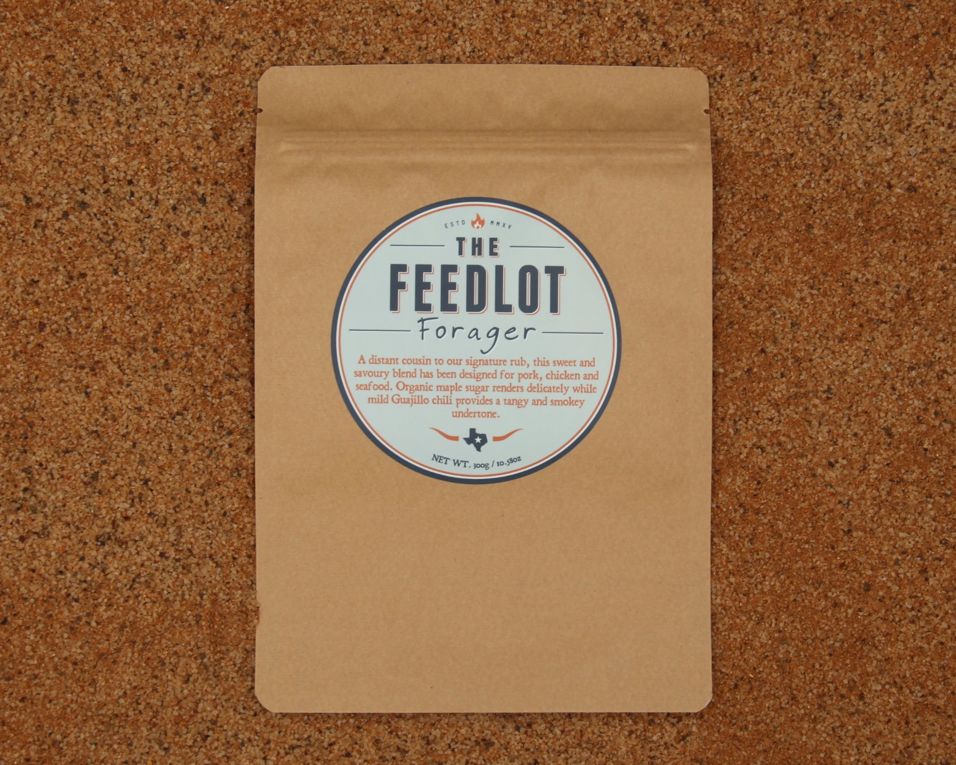 A brown paper package with a circular label reading “The Feedlot Forager.” The label describes the product as *Forager* by *The Feedlot*, a mild organic coffee sweetened with organic maple sugar and perfectly roasted for poultry, pork, and meaty dishes. The package is placed on a cork surface.