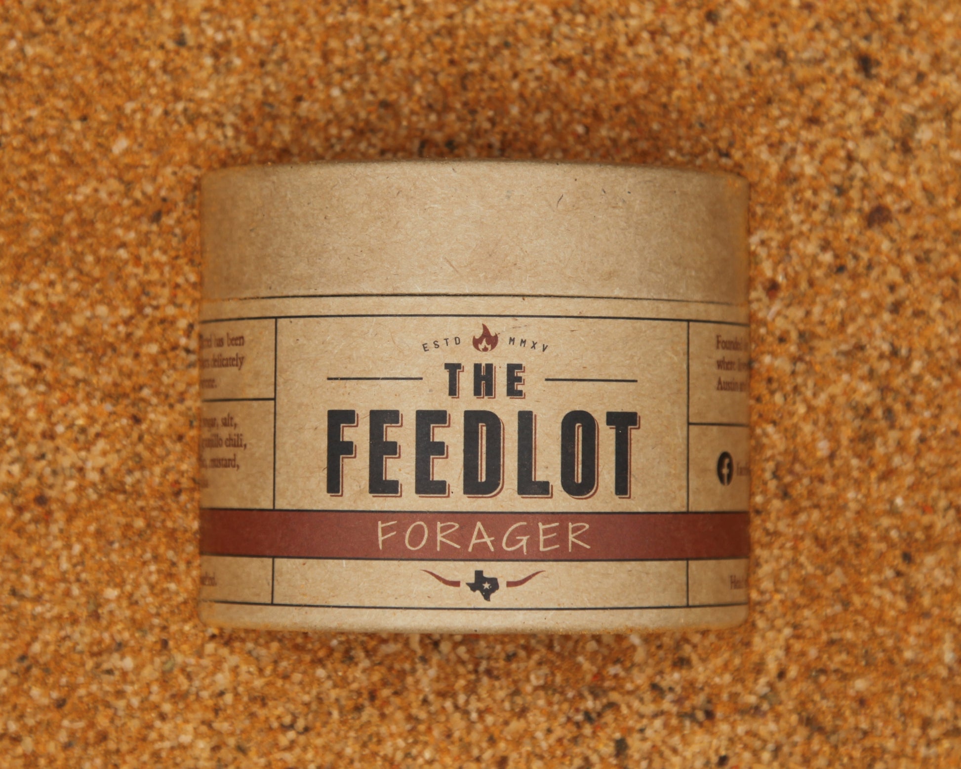 A cylindrical, brown paper container labeled "Forager" from The Feedlot sits on a textured, grainy surface. The label features bold black text with red accents and various smaller text details, including an established date and decorative elements. This blend includes organic maple sugar for added sweetness.