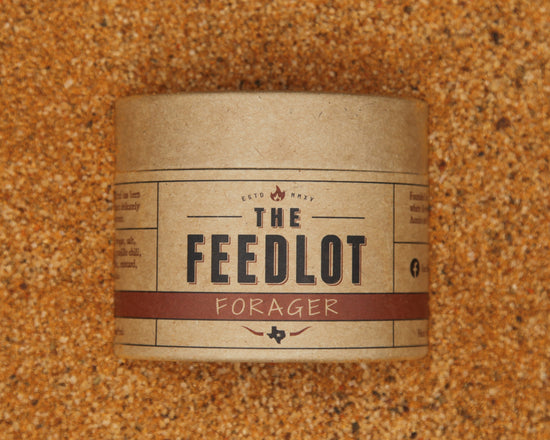 A cylindrical, brown paper container labeled "Forager" from The Feedlot sits on a textured, grainy surface. The label features bold black text with red accents and various smaller text details, including an established date and decorative elements. This blend includes organic maple sugar for added sweetness.