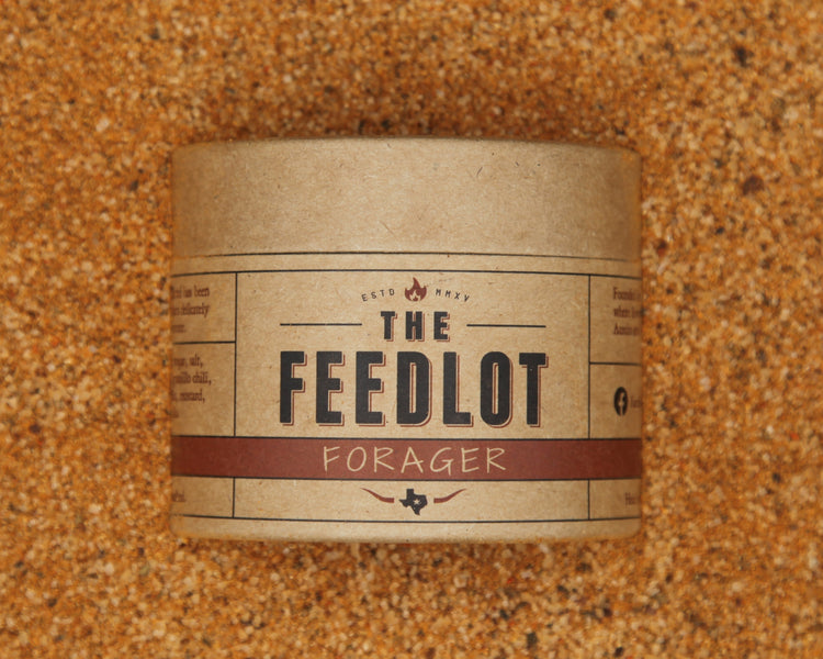 A cylindrical, brown paper container labeled "Forager" from The Feedlot sits on a textured, grainy surface. The label features bold black text with red accents and various smaller text details, including an established date and decorative elements. This blend includes organic maple sugar for added sweetness.