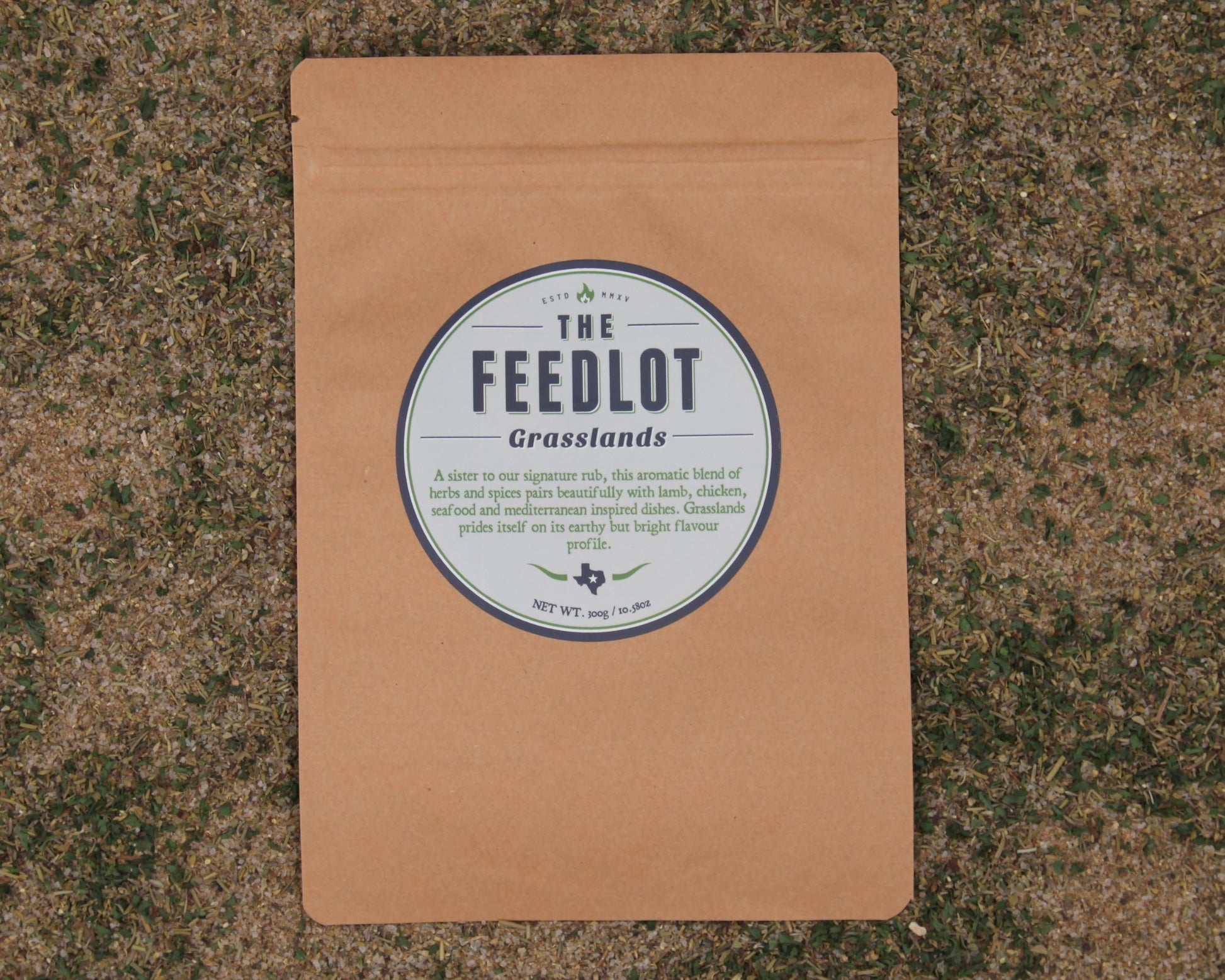 A brown paper package labeled "The Feedlot Grasslands" rests on grass. The label describes the blend as a mix of Mediterranean herbs and spices for beef, lamb, chicken, seafood, and vegetables. The package contains 240 grams or 8.46 ounces.