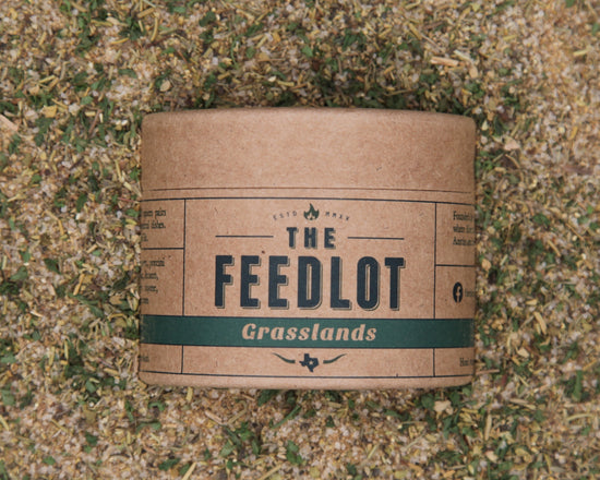 A brown cylindrical container with the label "Grasslands" by The Feedlot sits on a bed of mixed grass and herb fragments. The vintage design label, featuring smaller text, a flame icon, and other decorative elements, evokes rustic Mediterranean charm perfect for enhancing chicken or lamb dishes.