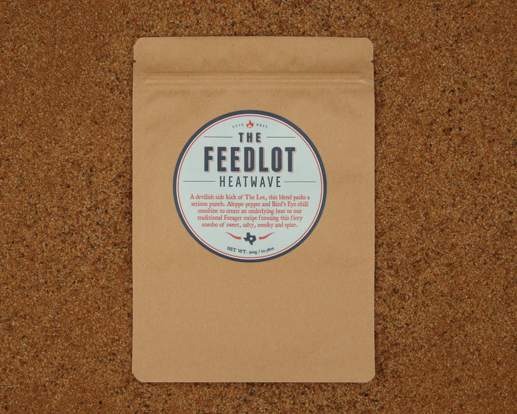 A brown paper package with a round label reading "Heatwave by The Feedlot." The label highlights a blend of spices, including ghost peppers and Aleppo pepper, describing the flavor as a combination of sweet and spicy with a smoky finish. The package rests on a cork surface.