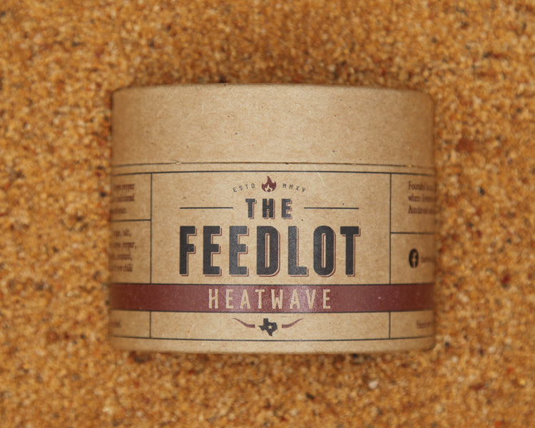 A cylindrical cardboard container labeled "Heatwave" from "The Feedlot" is centered in the image, placed on a textured brown background that resembles sand or finely ground spices. The branding, featuring Aleppo pepper and Bird's Eye chili, suggests it may contain a fiery food seasoning blend.