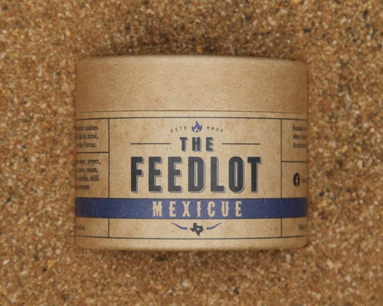 A cylindrical container labeled "The Feedlot Mexicue" rests on a sandy surface. The brown packaging with a blue band and black text hints at the Tex-Mex flavour inside, featuring small icons and details that evoke the rich tradition of Mexican food mixed with BBQ nuances.
